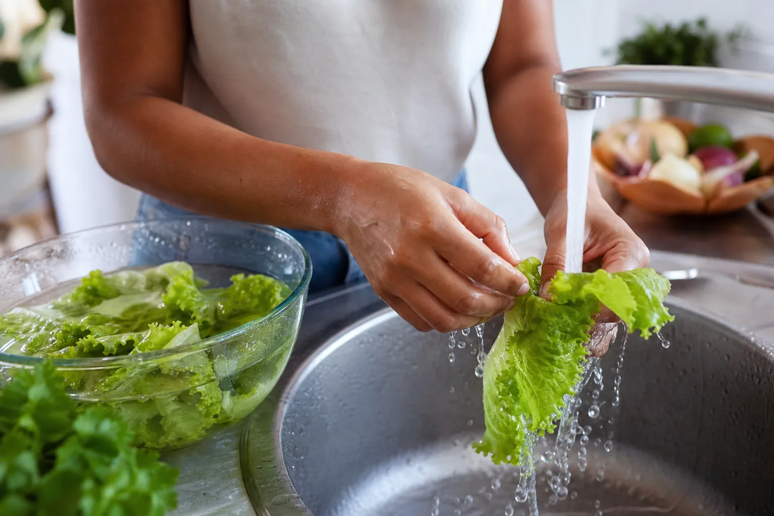 Mãos lavando folhas de alface uma a uma em água corrente na pia da cozinha