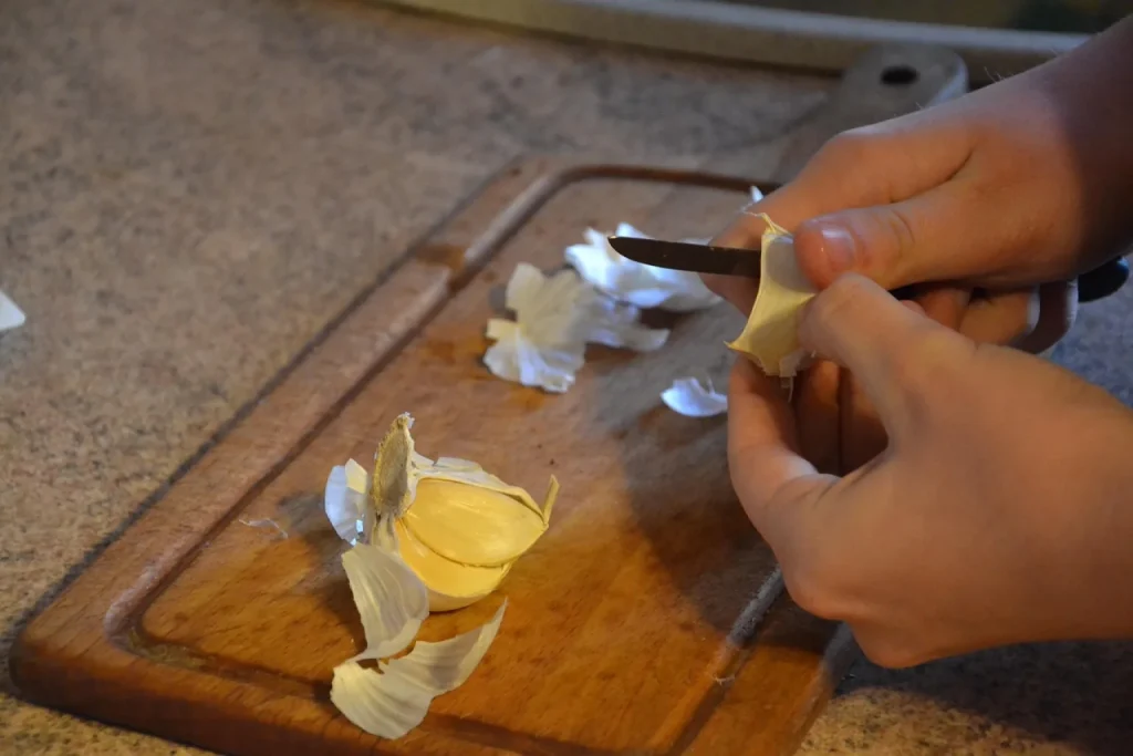 Mãos descascando dentes de alho sobre tábua de madeira, ao lado de panela de feijão verde, coentro e limão em cozinha de casa