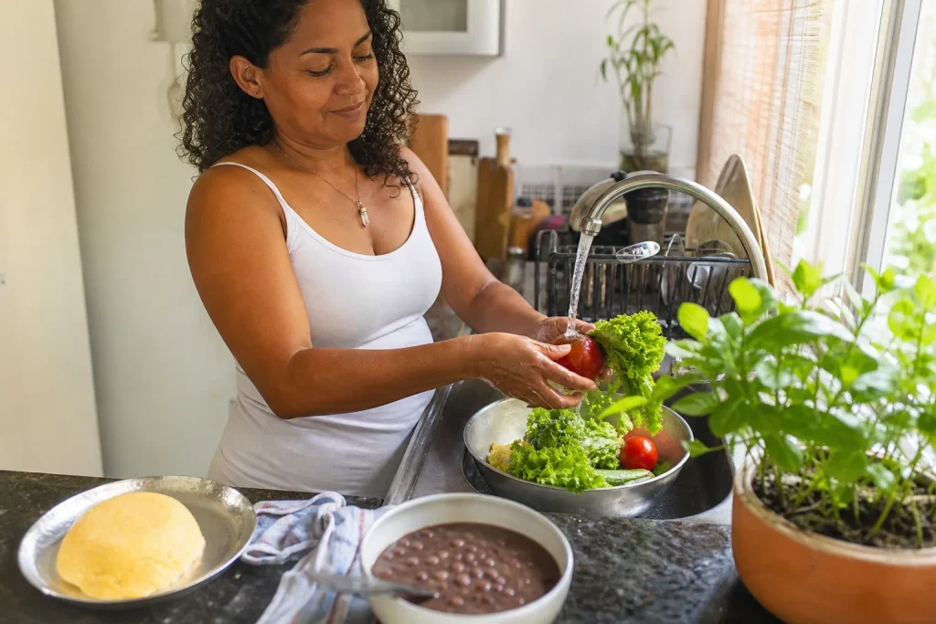 Mulher higienizando verduras e tomates na cozinha de casa, com alimentos brasileiros no balcão