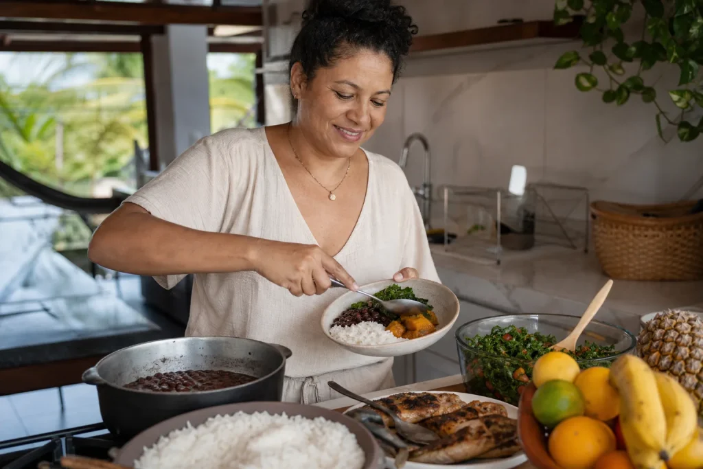 Mulher preparando prato brasileiro com feijão, arroz, verduras, peixe e frutas em cozinha de casa